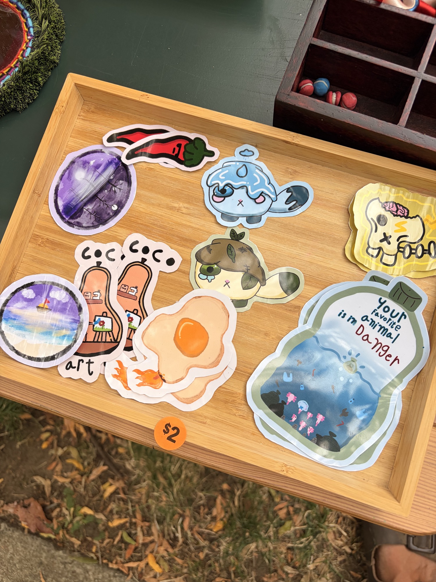 Hand-drawn stickers displayed on a wooden tray at the craft sale